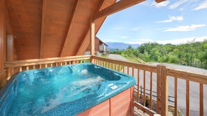 A wooden balcony with a hot tub overlooking a road and mountains.