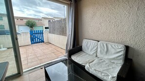 A room with a sliding glass door, a white sofa, and a view of a residential area with a blue gate. - Cozy 2-Room House with Mezzanine, Pool & Parking - Saint-Cyprien Beach (Saint-Cyprien)