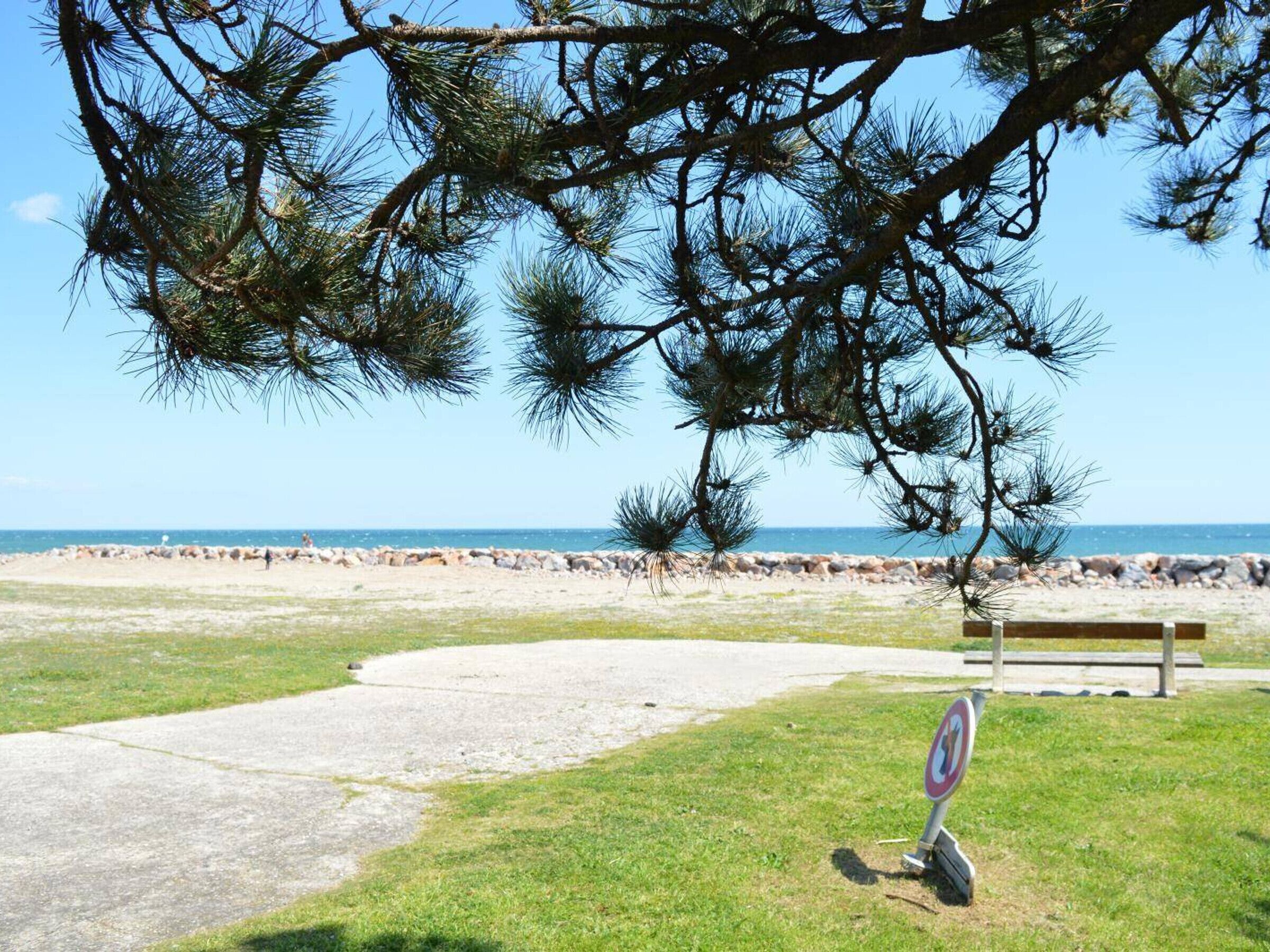Ein gepflasterter Weg führt zu einer Bank an einem Strand, der von einem klaren Himmel überdacht wird.