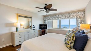 Um quarto com cama, cômoda, ventilador de teto, televisão e janela com vista para o oceano.
