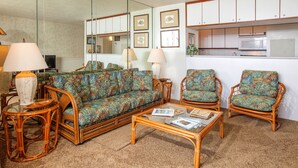 Una sala de estar con sofá, dos sillones, una mesa de centro, televisor y una zona de cocina con armarios y microondas.
