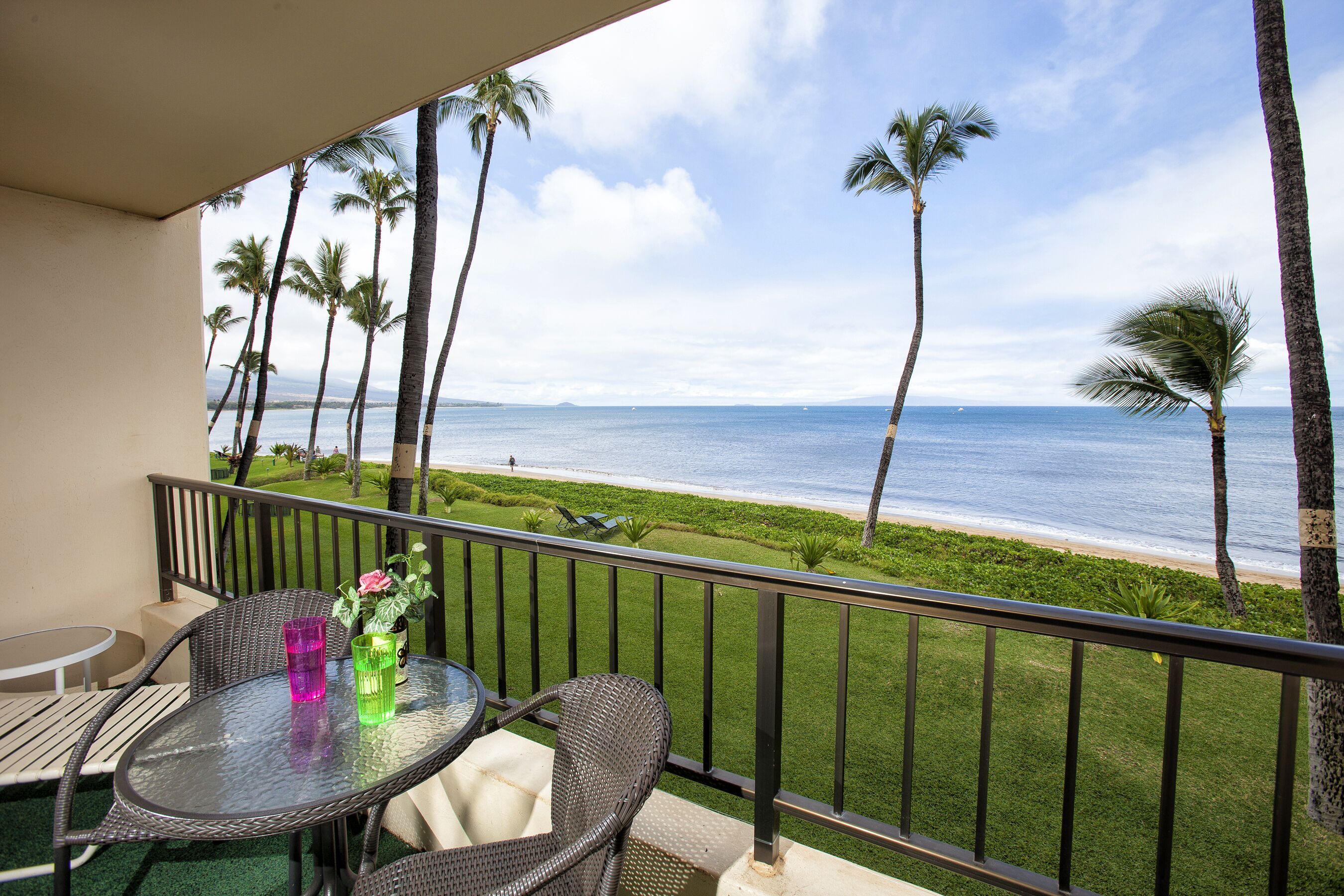 A balcony with a glass table, wicker chairs, and a view of the beach and ocean.