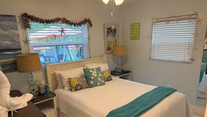 A neatly arranged bedroom with a bed, bedside tables, a window with blinds, and a view of a balcony. - Castaway Cottage + Beach + Heated Private Pool & Spa + GOLF CART INCLUDED! (Anna Maria)