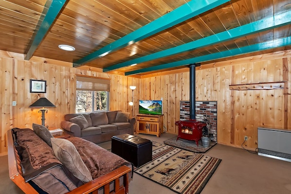 A cozy log cabin interior with a wooden ceiling, a fireplace, a sofa, a television, and a window with a view of trees.