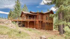 A large, rustic log cabin with a balcony and multiple windows, surrounded by pine trees and a stone wall.
