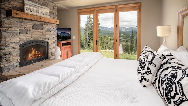Un dormitorio con chimenea de piedra, una cama con ropa de cama blanca, vistas a montañas a través de grandes ventanales y un televisor sobre un estante de madera.