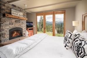 A bedroom with a stone fireplace, a bed with white bedding, a view of mountains through large windows, and a television on a wooden shelf. - Cowboy-Chic, Ski-In/Ski-Out Home with Mountain Views! (Big Sky)