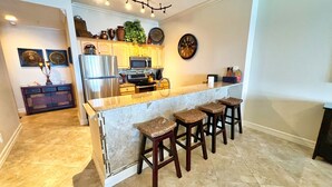 A modern kitchen with a marble island, bar stools, stainless steel appliances, and decorative items on the wall. - 4 Bedroom Platinum Designer End Unit Incredible Gulf Views! Free Beach Chairs (Panama City Beach)