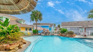 A swimming pool with a rock feature, surrounded by colorful buildings and palm trees. - Spacious coastal cottage at Pirates Bay! Just a short drive to the beach and one (Port Aransas)