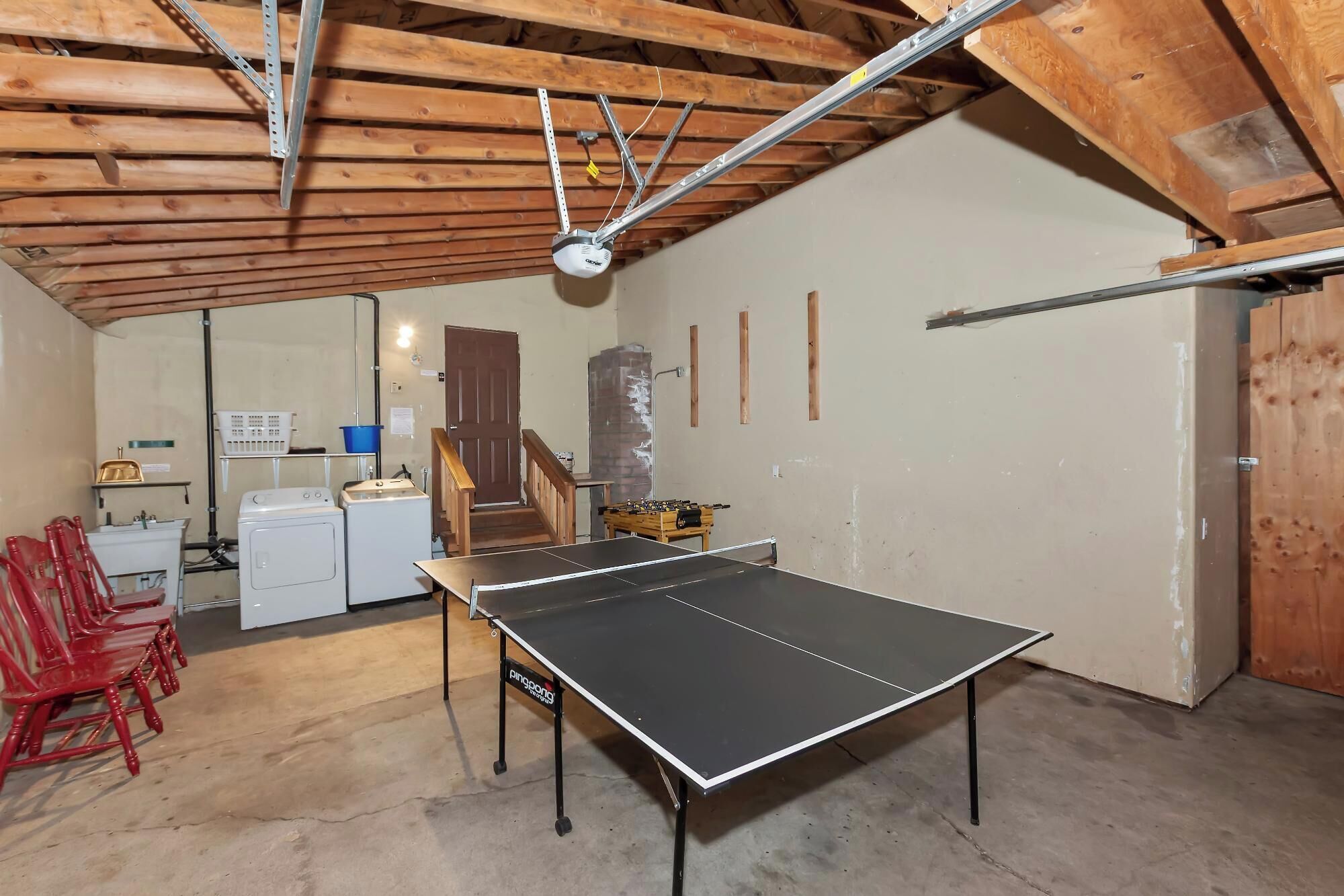 Een tafeltennistafel in een kamer met houten balken, een wasmachine en rode stoelen.