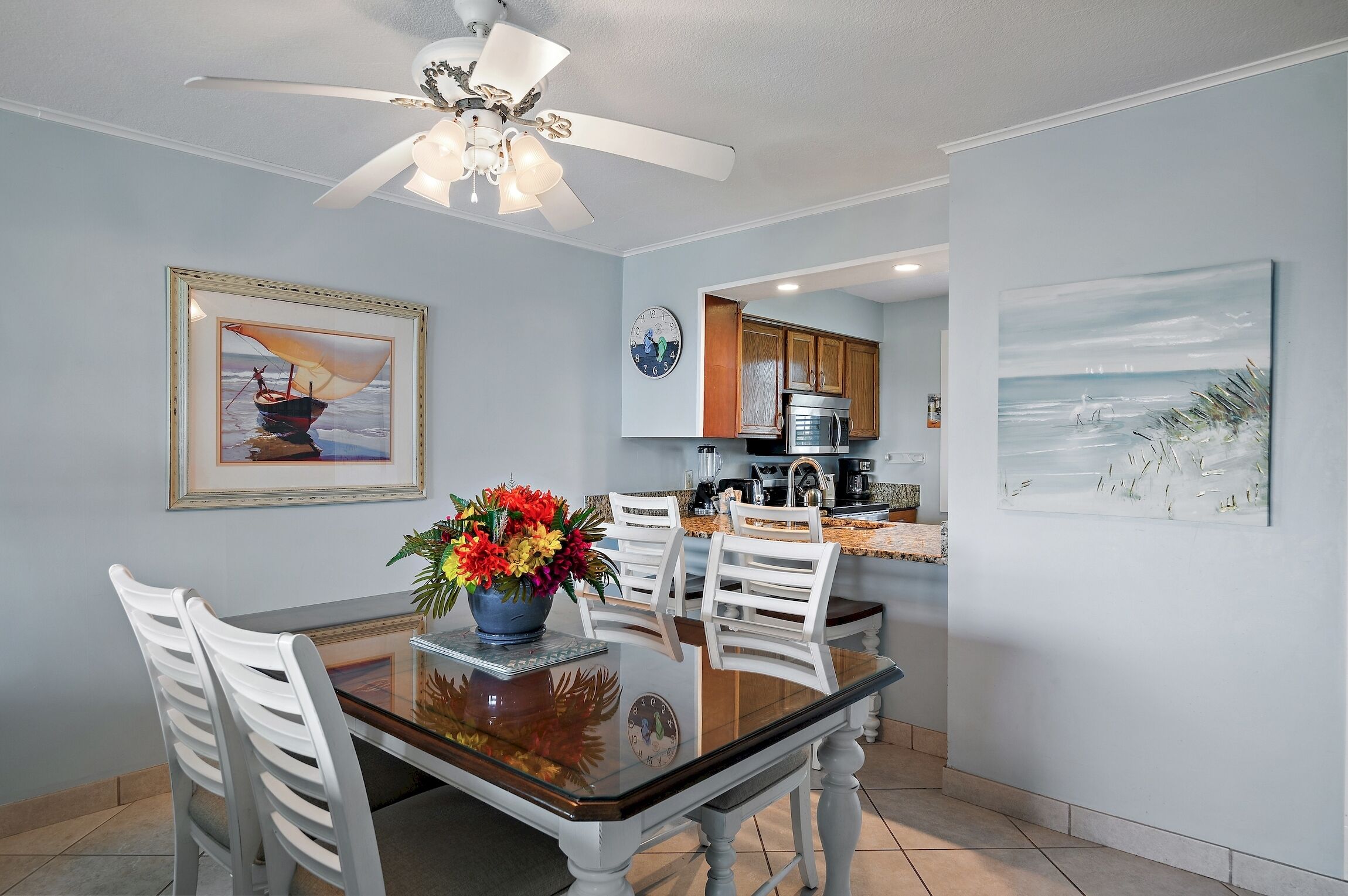 A dining area with a glass table, white chairs, a ceiling fan, and artwork on the walls.