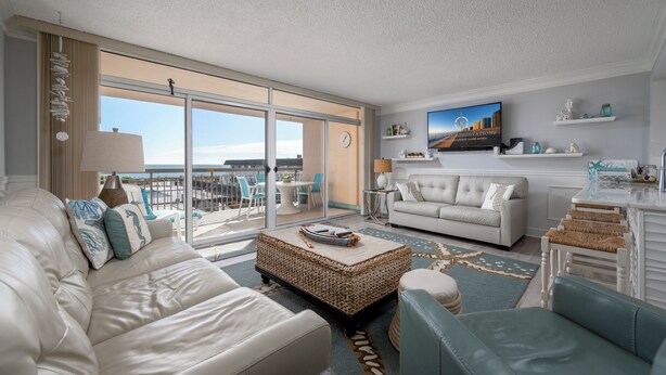 A modern living room with a white sofa, a blue armchair, a wicker coffee table, and a television mounted on the wall. - Beautiful one bedroom oceanfront condo with stunning ocean views and a pool! (Ocean City)
