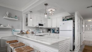 A modern kitchen with white cabinetry, a large island with stools, stainless steel appliances, and decorative items on shelves. - Beautiful one bedroom oceanfront condo with stunning ocean views and a pool! (Ocean City)
