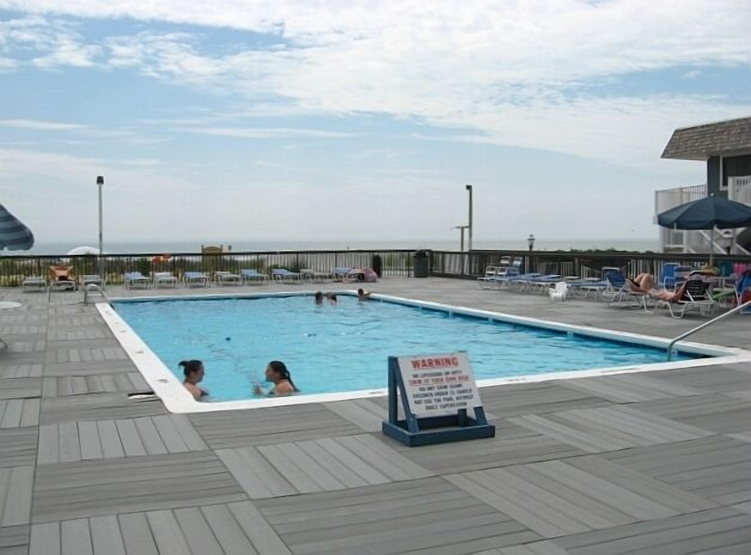 A pool area with a warning sign, lounge chairs, and a view of the ocean.
