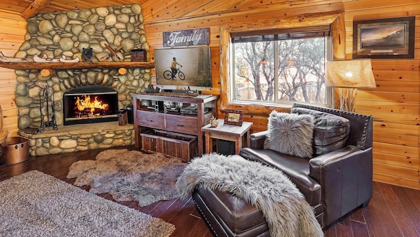 A cozy cabin interior with a stone fireplace, leather furniture, and a large window offering a view of trees.