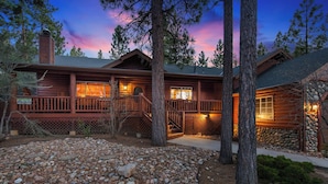 A log cabin with a wooden deck, stone foundation, and a view of the surrounding forest.
