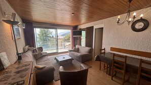 A cozy living room with a sofa, armchair, and coffee table. There is a dining table with chairs and a chandelier. The room has a wooden ceiling and a large window with a view of the outdoors. - Spacious 4-Room Apartment for 9 - Ski Lift, Balcony, Garage - Alpe d'Huez (Huez)