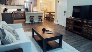 A modern kitchen with stainless steel appliances, a dining area with bar stools, a living room with a sofa and coffee table, and a TV on a wooden cabinet.
