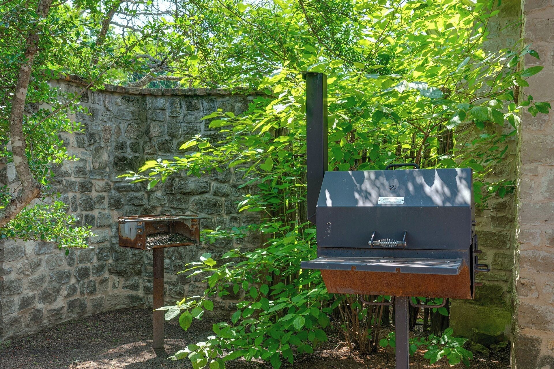 Una parrilla con chimenea, un contenedor metálico oxidado y un muro de piedra al fondo.