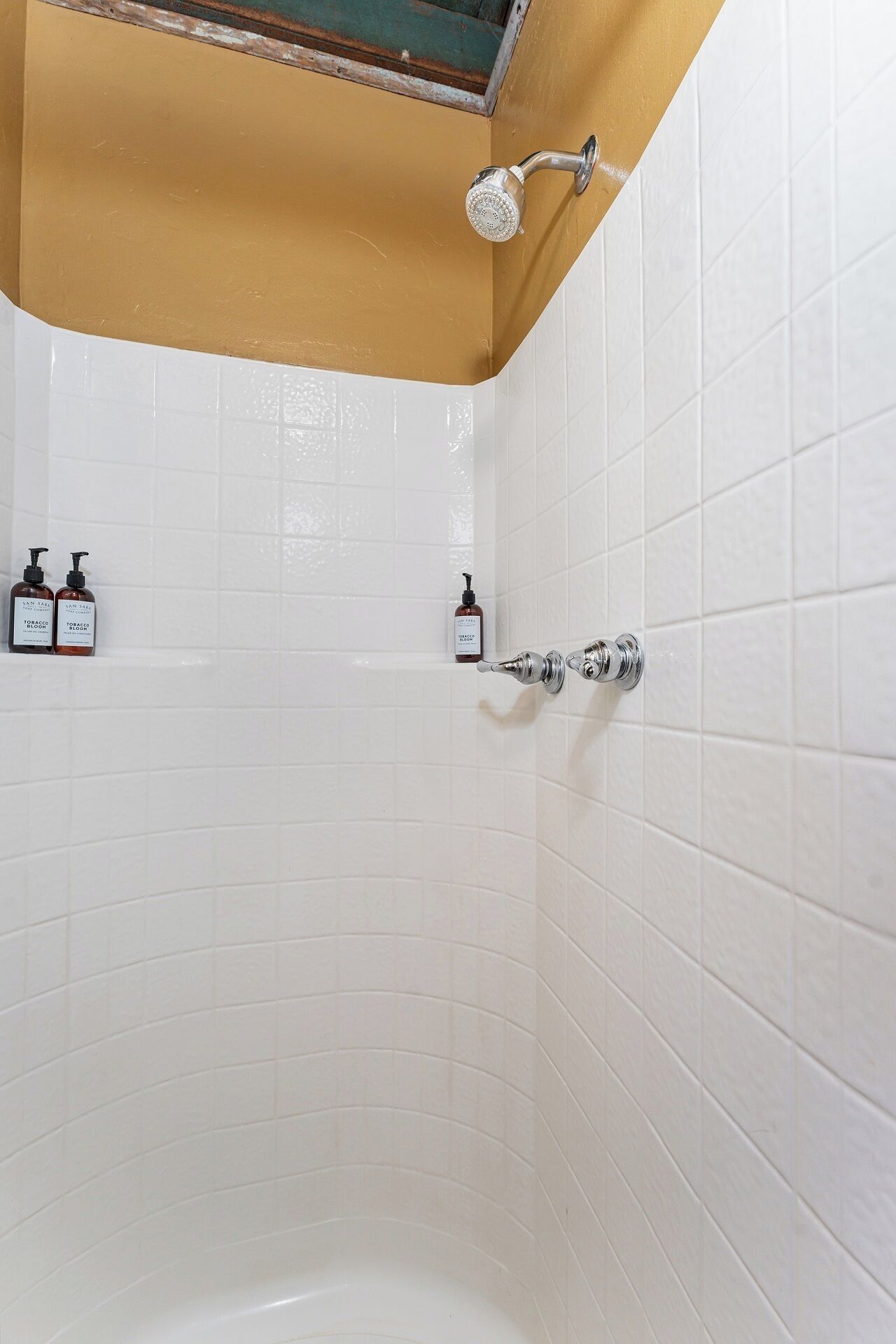 Un bagno con pareti piastrellate di bianco, una doccia con soffione manuale e due flaconi di articoli da toilette a parete.