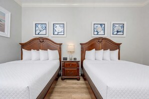 Una habitación de hotel con dos camas de madera, ropa de cama blanca, una mesita de noche con lámpara y cuadros en la pared.
