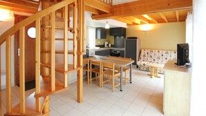 A wooden staircase leading to a loft bedroom, a kitchen with modern appliances, and a living area with a sofa and a TV. - Cozy house in La Hague (La Hague)