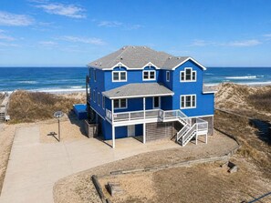 A two-story blue house with a balcony and a staircase leading to the second floor, located near a beach with the ocean in the background. - Ocean Dream - Corolla, Ocean Sands, Pool/Hot Tub/Handicap Friendly/Elevator/Pets OK/Weddings! (Corolla)