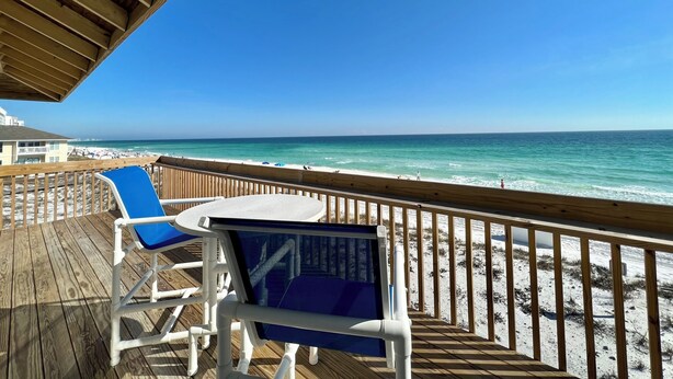 A wooden deck with a view of the ocean, a white table, and blue chairs.