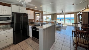 A kitchen with a refrigerator, microwave, and stove, a dining area with a table and chairs, and a balcony with a view of the ocean.