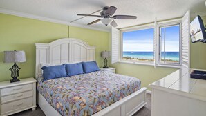 Une chambre à coucher comprenant un lit, des tables de chevet, un ventilateur de plafond, une fenêtre donnant sur la plage et un bureau.