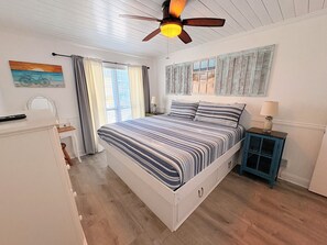 Ein Schlafzimmer mit einem Bett, einem Deckenventilator, einem Fenster mit Vorhängen, einem Nachttisch und einer Wandlampe.