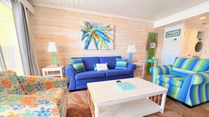 Ein gemütliches Wohnzimmer mit einem blauen Sofa, einem gestreiften Sessel und einem blumigen Sessel, einem Couchtisch und einem Wandbild.