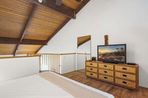 Une chambre avec un plafond en bois, un lit, un téléviseur posé sur une commode en bois, et une vue sur la plage par la fenêtre.