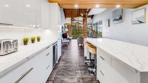 A modern kitchen with white cabinets, a wooden ceiling, and a view to a living area with a staircase. - Roomy Maui Suite w/Loft, Relaxing Lanai, Kitchen Ease, WiFi–Kamaole Sands 10408 (KIHEI)