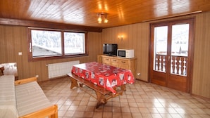Una habitación con paneles de madera, una cama, una mesa con mantel rojo, un televisor y vistas a montañas nevadas a través de amplios ventanales.