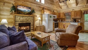 A cozy log cabin interior with a stone fireplace, wooden furniture, and a kitchen area.