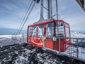 Um funicular vermelho com um logotipo triangular, suspenso por cabos, com vista para uma paisagem montanhosa coberta de neve.