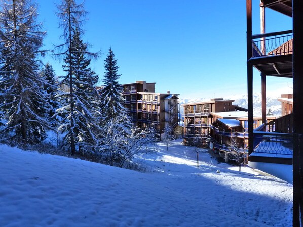 A snow-covered landscape with modern buildings, including a multi-story structure with balconies and a wooden cabin.