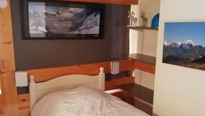A bedroom with a bed, a mounted TV, a wooden shelf with a moose decoration, and a framed picture of mountains.