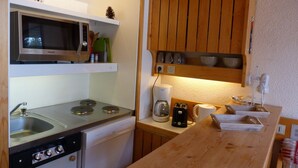 A compact kitchen with a microwave, stove, and sink. There are wooden cabinets above the counter and a shelf with glassware.