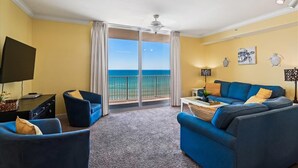 A living room with a blue sofa, a TV, and a balcony with a view of the ocean. - Tidewater 1 bedroom with gulf view, Summer dates open! (Panama City Beach)