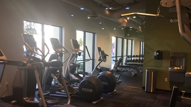 Un gimnasio con diferentes equipos de ejercicio, ventanas amplias y un dispensador de agua.