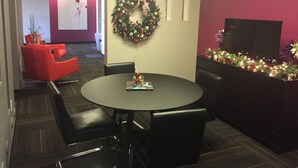 Un área de recepción con una mesa redonda, sillas negras, un sofá rojo, una corona navideña y un televisor sobre un mueble.