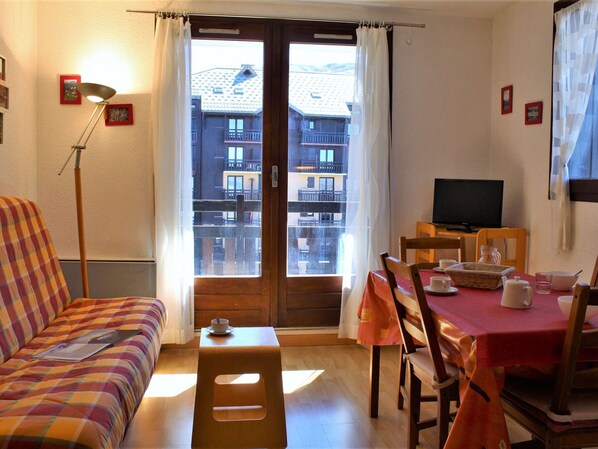 A cozy room with a sofa, a dining table with chairs, a television, and a view of buildings through the window.
