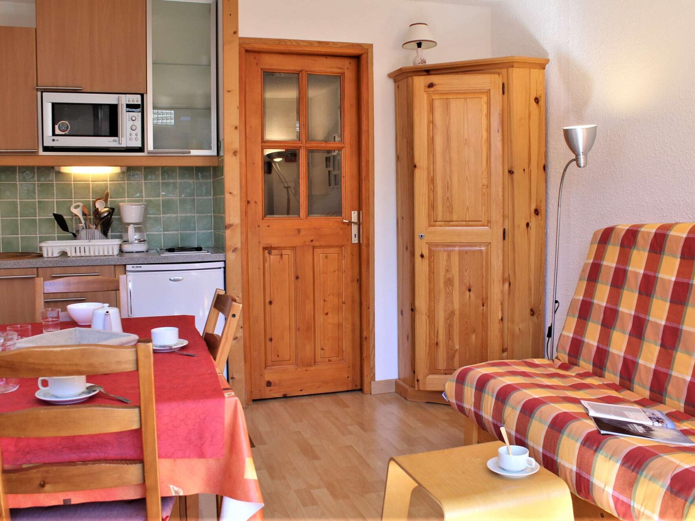 A compact kitchen with a microwave, a coffee maker, and a dining table with chairs. There is a wooden cabinet and a plaid sofa in the living area.