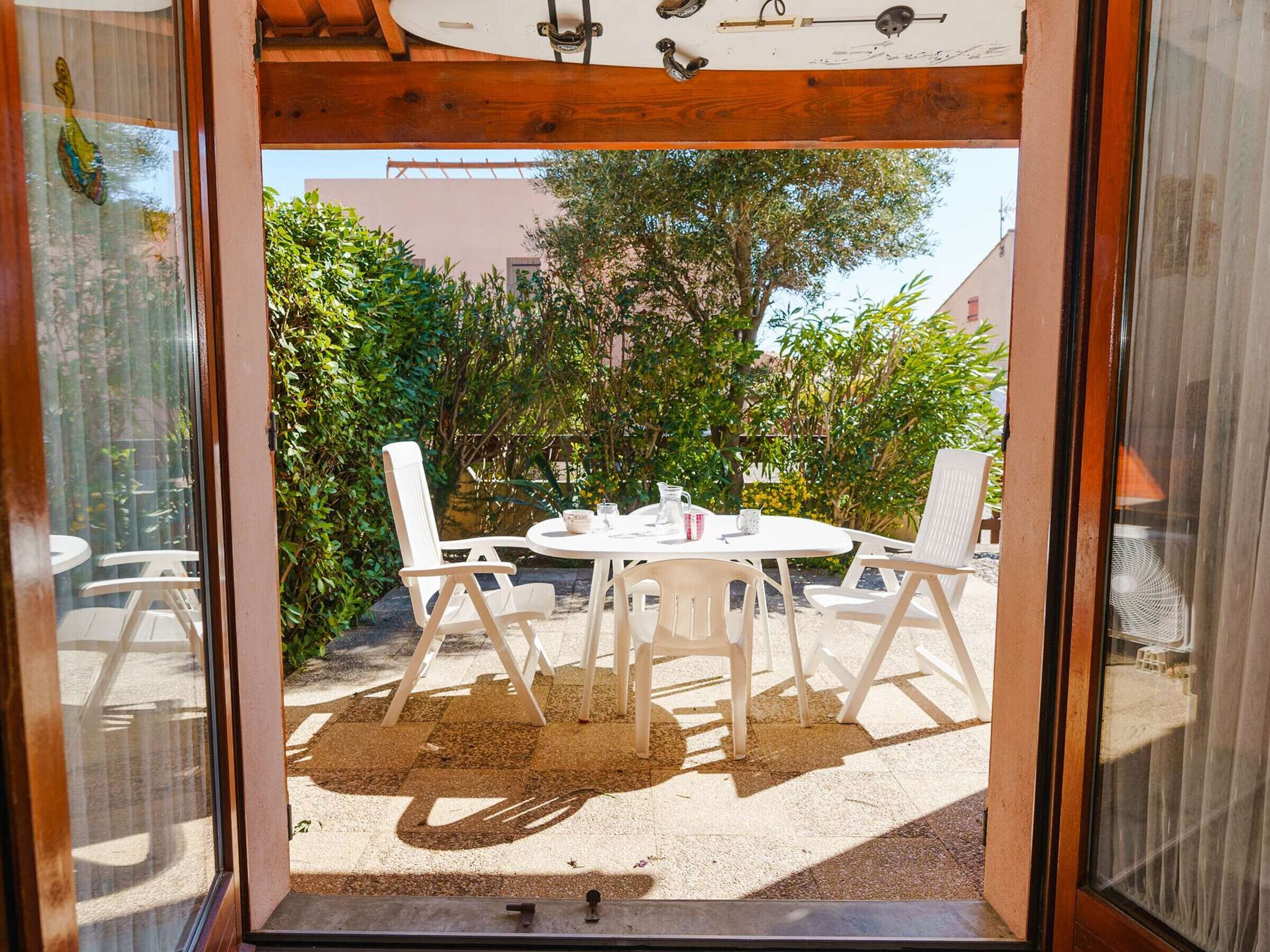 A patio with a round table and four chairs, surrounded by greenery and a building in the background.