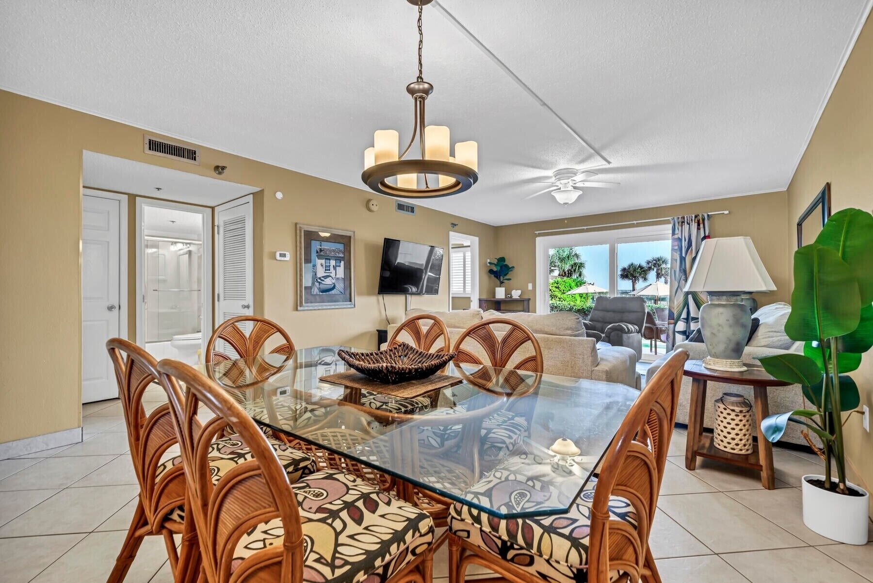 A spacious dining area with a glass-top table surrounded by wicker chairs, a ceiling fan, and a television mounted on the wall.
