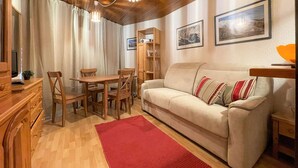 A cozy living room with a wooden ceiling, a beige sofa, a red rug, a wooden dining table with chairs, and a bookshelf.