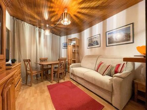 Uma sala de estar acolhedora com teto de madeira, um sofá bege, um tapete vermelho, uma mesa de jantar de madeira com cadeiras e uma estante de livros.