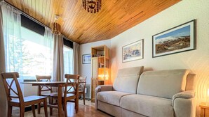 A cozy living room with a wooden ceiling, a beige sofa, wooden chairs, a dining table, and framed pictures on the wall.